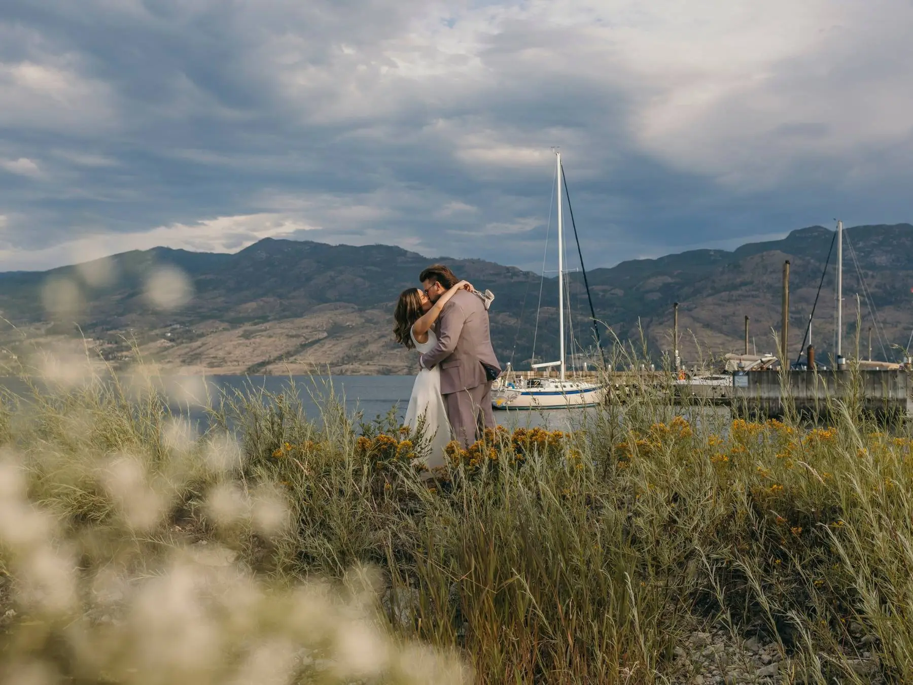 A bride and groom kissing among wildflowers beside a marina, mountains and stormy skies behind them