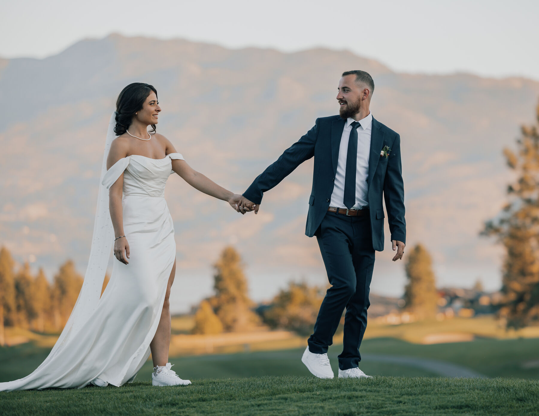 Bride in a white gown walking hand in hand with the groom on a golf course at sunset