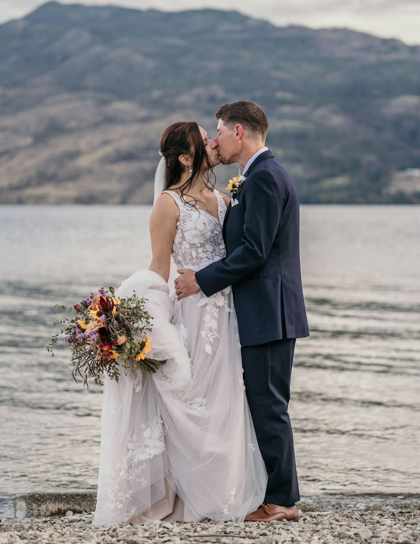Bride and groom kissing on a pebble beach, the hills behind them
