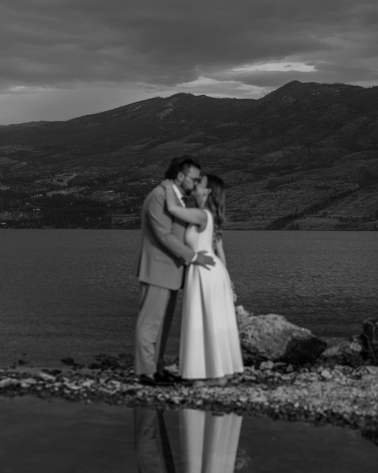 Black and white photo of a couple embracing on a rocky shore with their reflection in the water